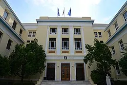 Image of a yellow building with trees in the forefront and three flags on the top of the building. 